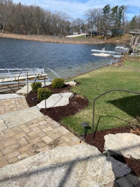 Stone steps descend through a landscaped yard with mulch and shrubs toward a lake featuring boat docks and a blue sky.