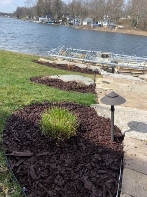 A view of a grassy shoreline with dark mulch garden beds, a stone pathway, a metal dock structure, and a lake in the back.