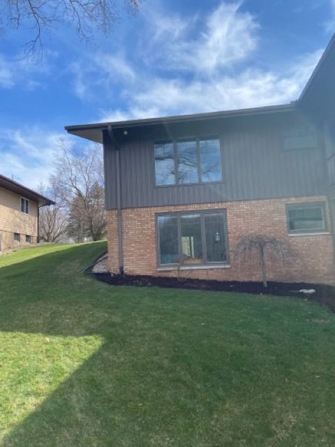 Two-story house with brown vertical siding, brick lower level, gray windows, and a sloping lawn under a clear blue sky.