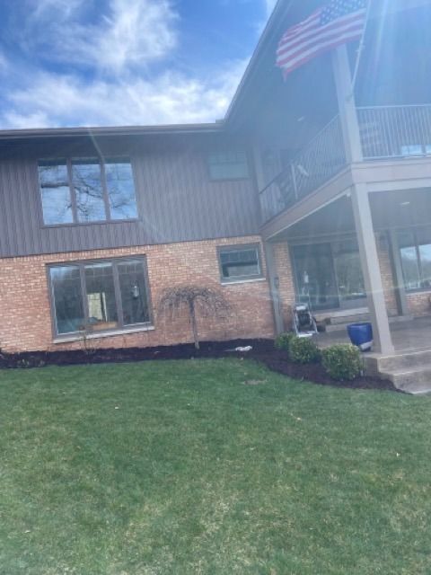 A two-story home with tan brick, gray siding, a small weeping tree, and a second-story balcony with an American flag.