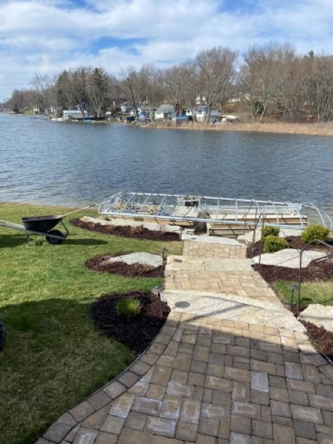 A stone walkway leads toward a metal dock on a lake, with green lawns, landscaped mulch beds, and a wheelbarrow nearby.