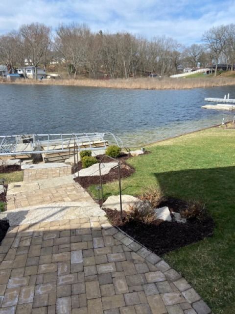 A stone paver walkway leads down a grassy slope toward a lake with a metal dock in the water on a sunny day.