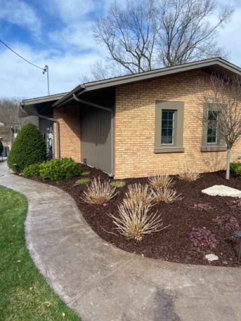 A tan brick building with two windows and dark brown trim, situated next to a curved concrete path and mulched landscaping.