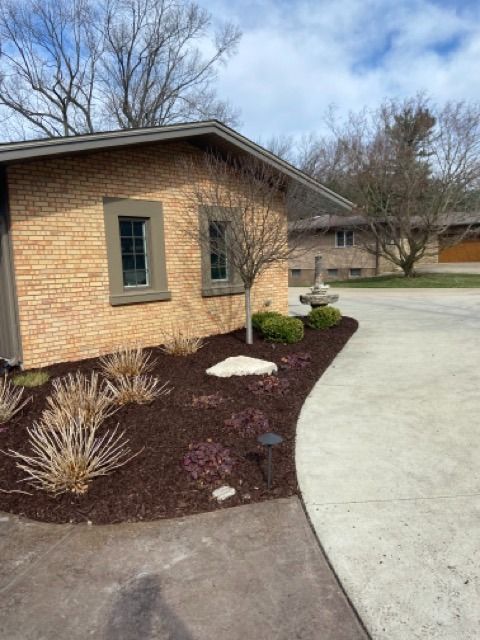 A light tan brick building with two windows, surrounded by a mulch bed with small plants and a concrete driveway.