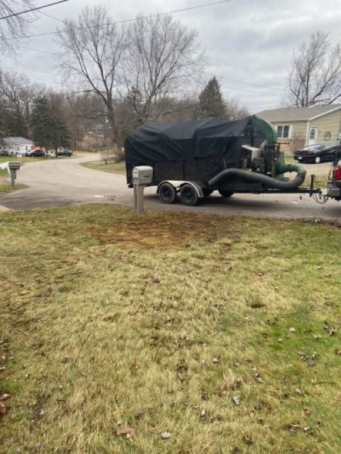 A black leaf vacuum trailer parked on a suburban street in front of a residential lawn.