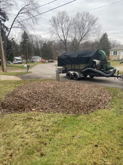 A large pile of dry leaves sits on a grass lawn next to a street, with a leaf vacuum trailer parked nearby.
