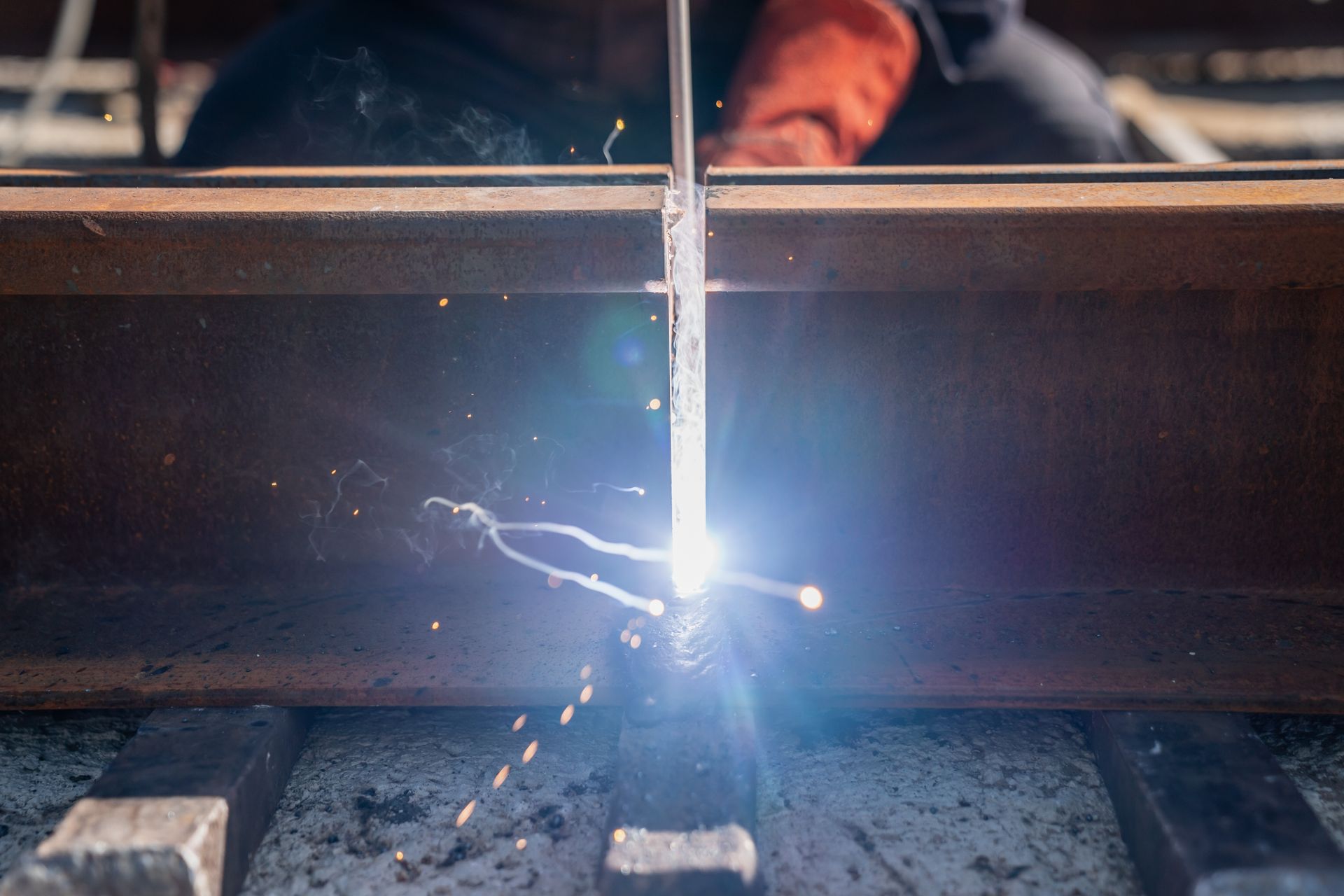 Welding sparks fly as a worker joins metal beams in a dim workshop.