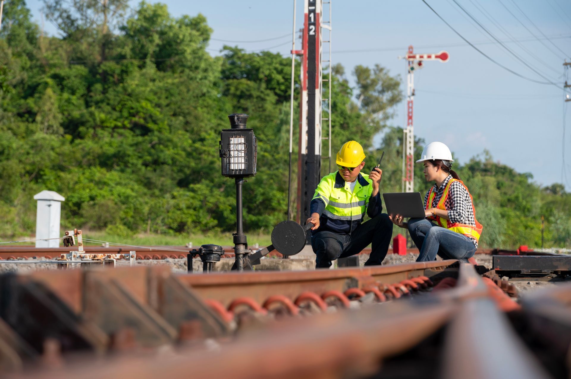 Two railway workers in hard hats inspect tracks outdoors near a signal and trees.