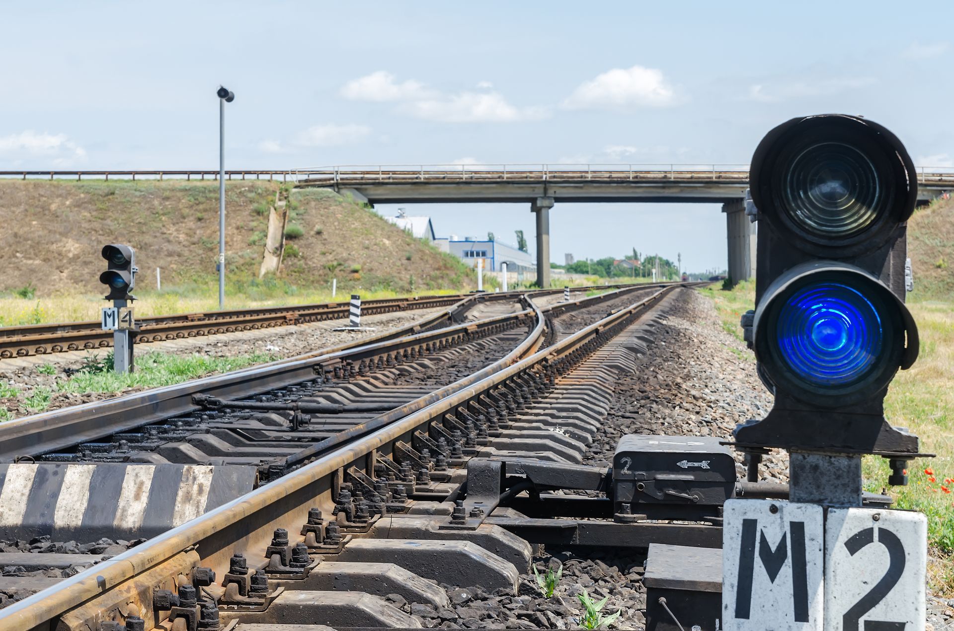 Railroad tracks under an overpass, with a blue signal light and M2 sign in the foreground.