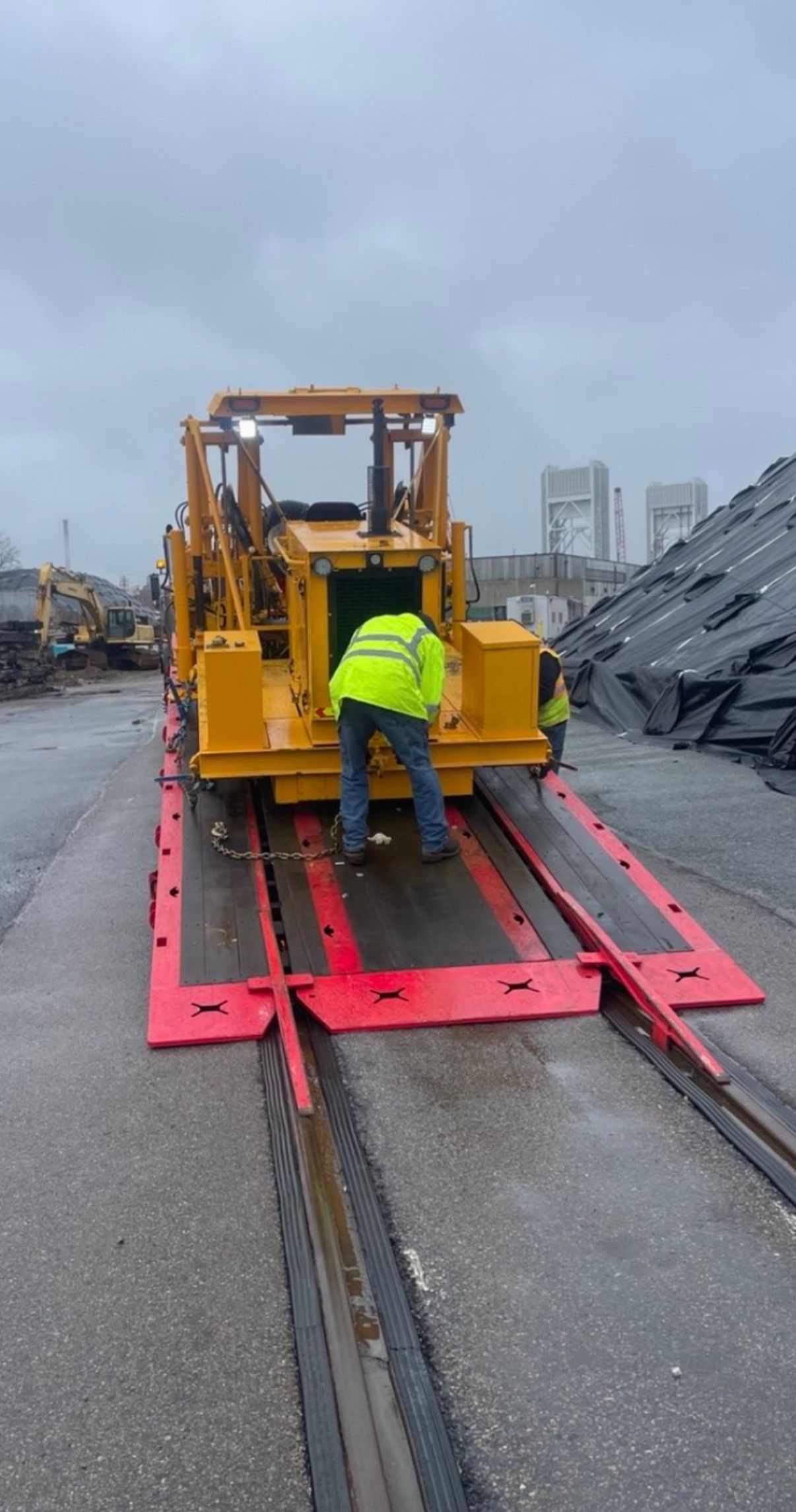 Worker in neon jacket inspecting a yellow machine on red rails at a rainy industrial site.