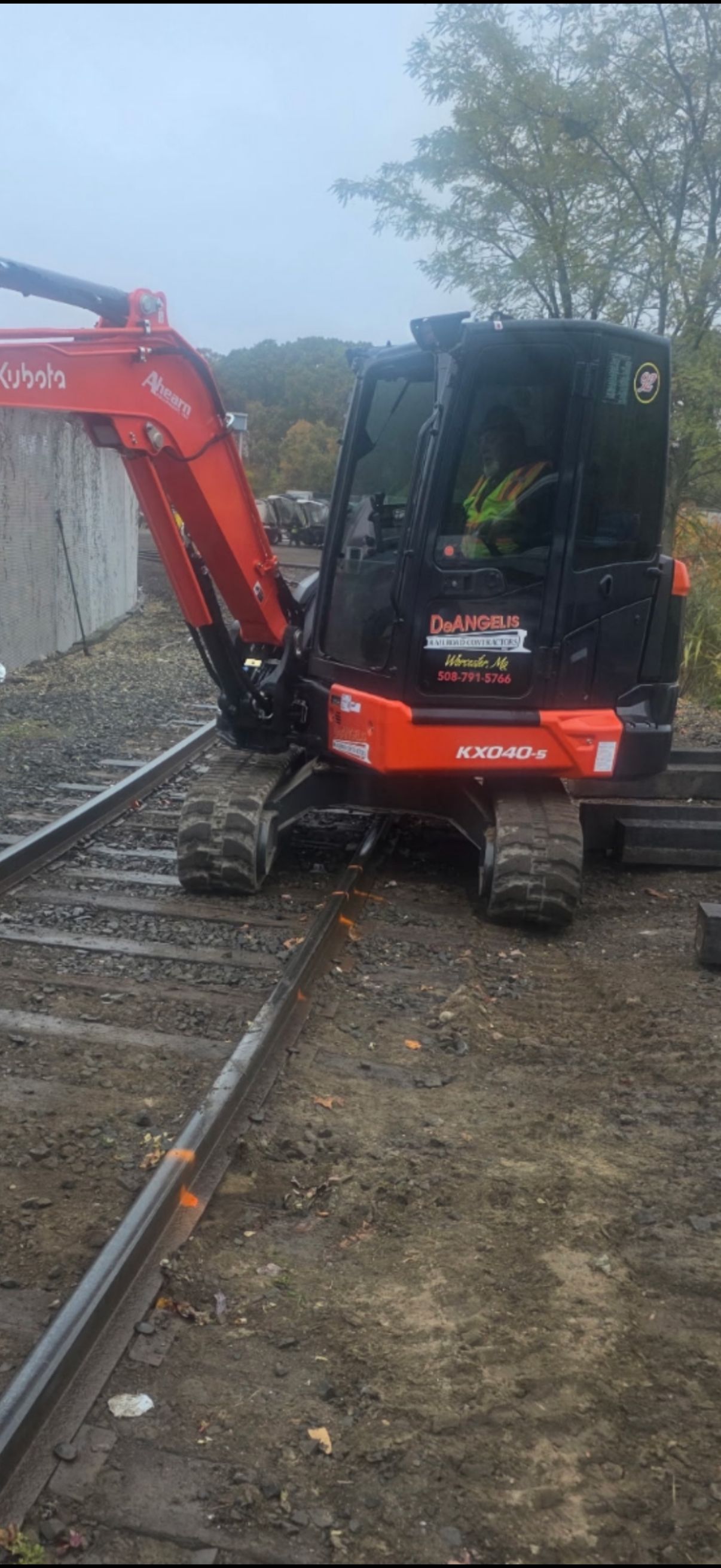 Orange excavator beside railroad tracks on a wet gravel worksite, with a black cab and boom extended.