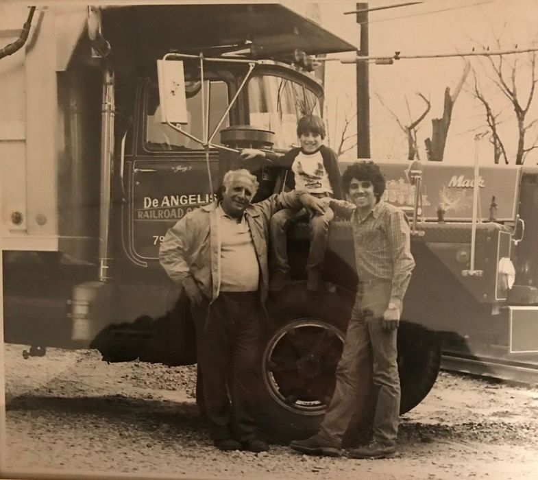 Three people pose beside a vintage truck, with one child sitting on the large front tire.