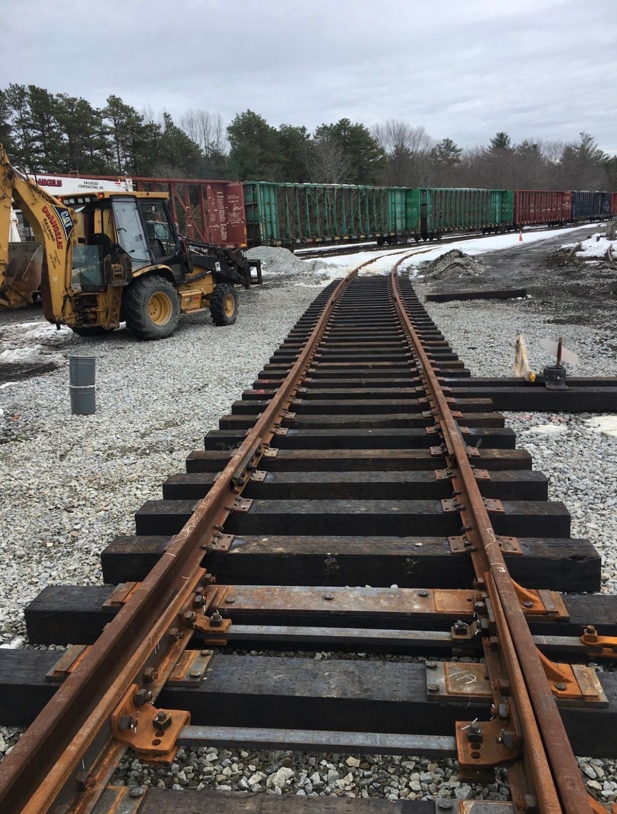 Railroad tracks in a snowy yard with a backhoe and freight cars in the background.