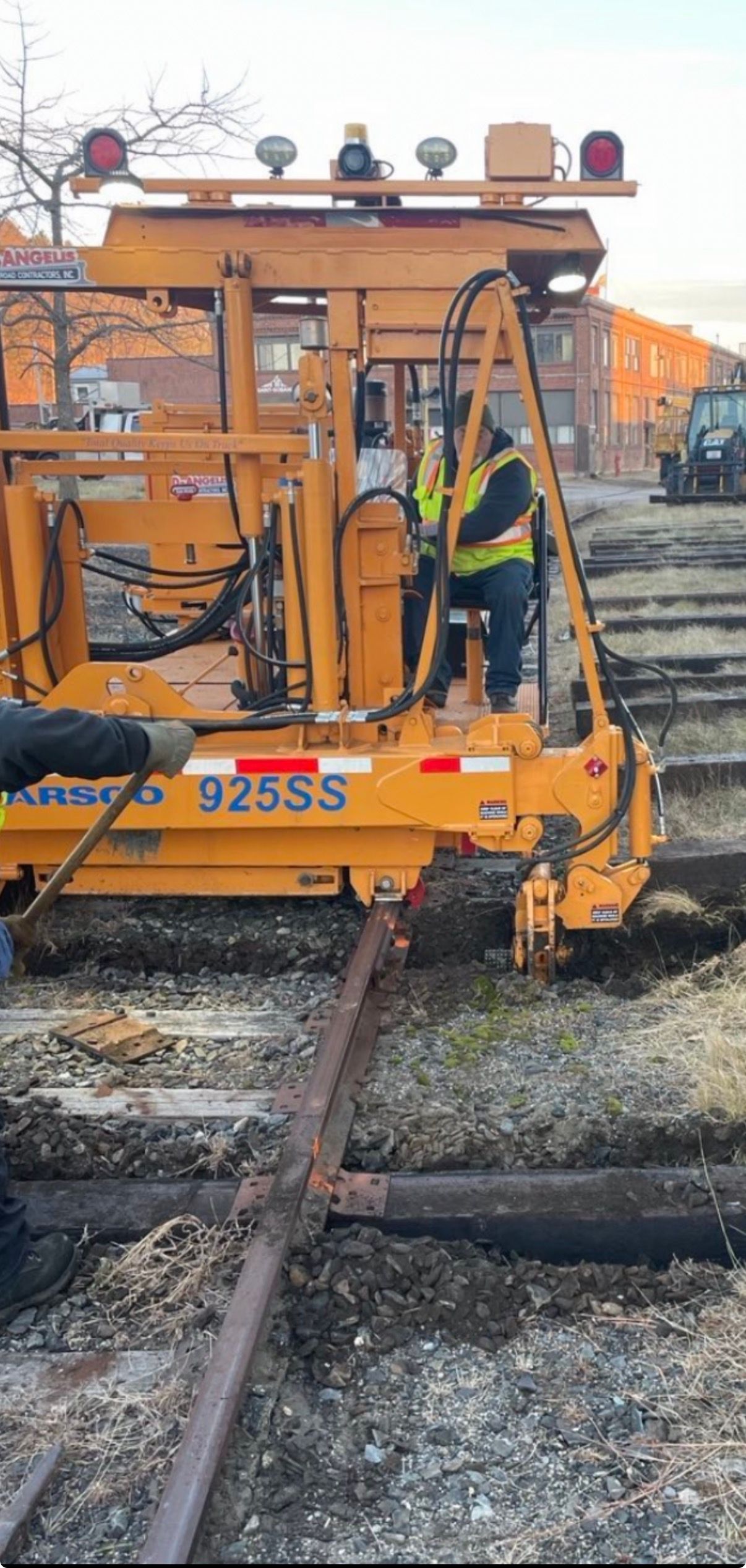 Yellow rail maintenance machine on train tracks in a gravel yard, with hoses and tools attached.