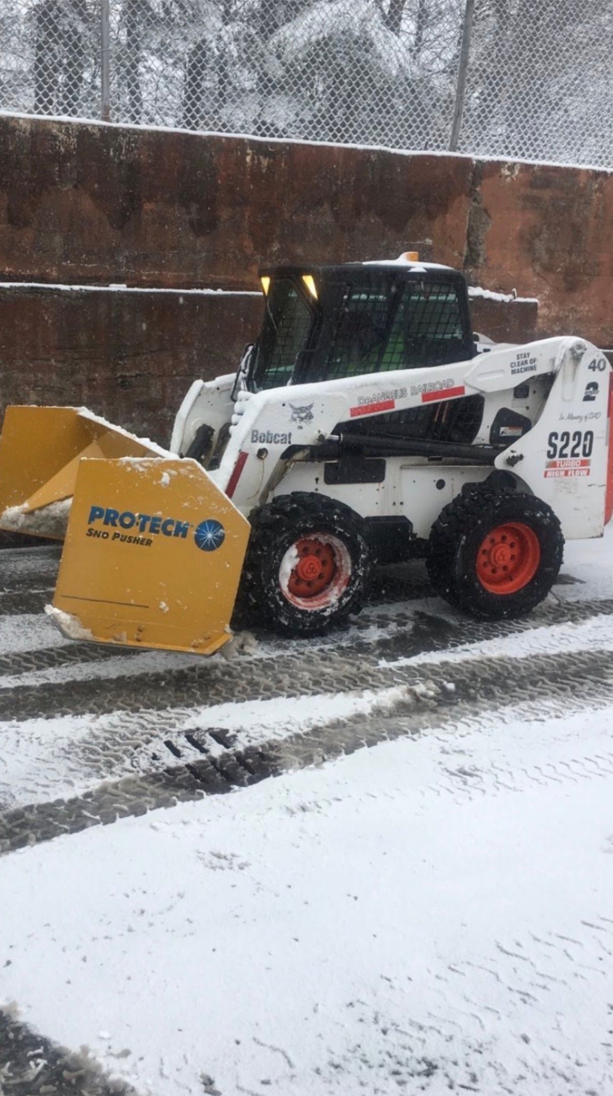 White skid-steer loader pushing snow with yellow blade on a snowy street beside a brick wall