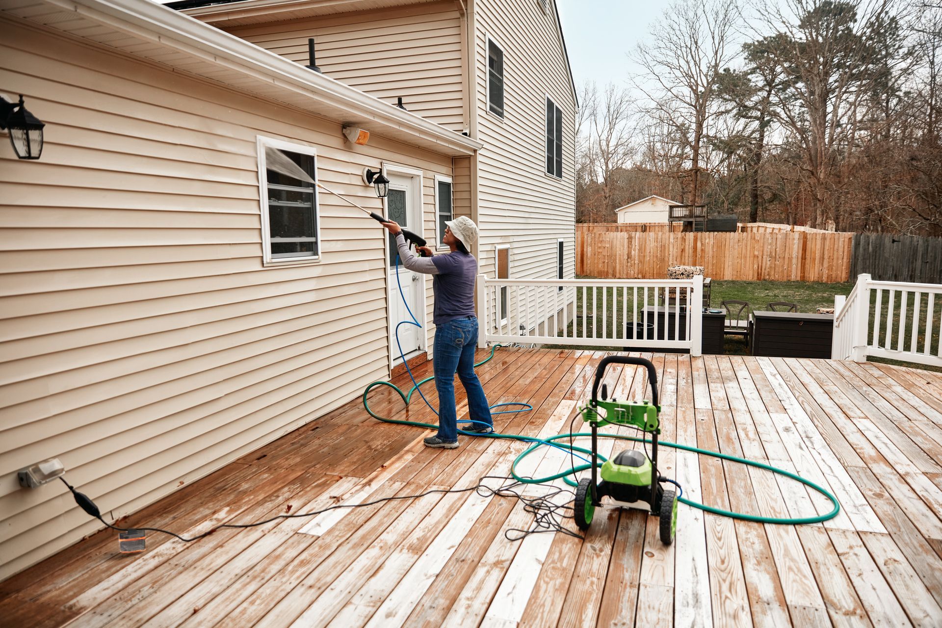 A person power washing a wooden deck attached to a light tan house on a cloudy day. A person power washing a wooden deck attached to a light tan house on a cloudy day.