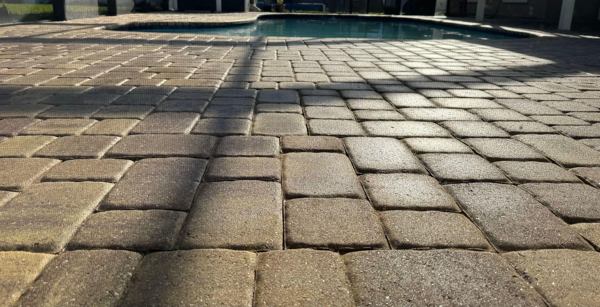 Brick patio around a swimming pool, tan and gray bricks, sunny outdoor setting.