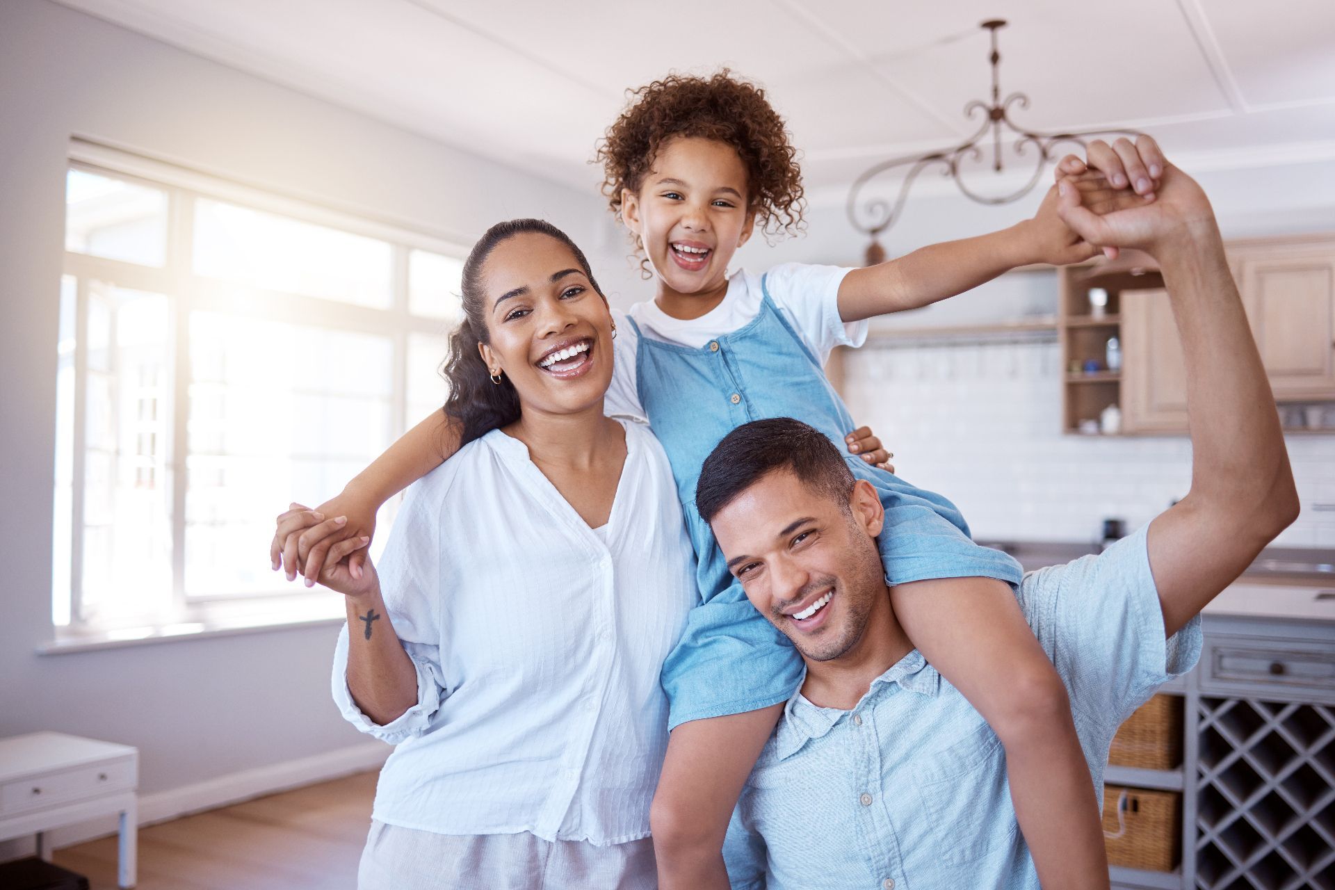 Smiling family in a bright living room, child on father's shoulders while mother stands beside them.