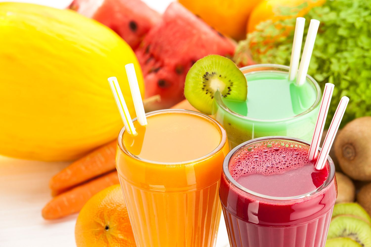Three different colored smoothies with straws and fruit in the background.