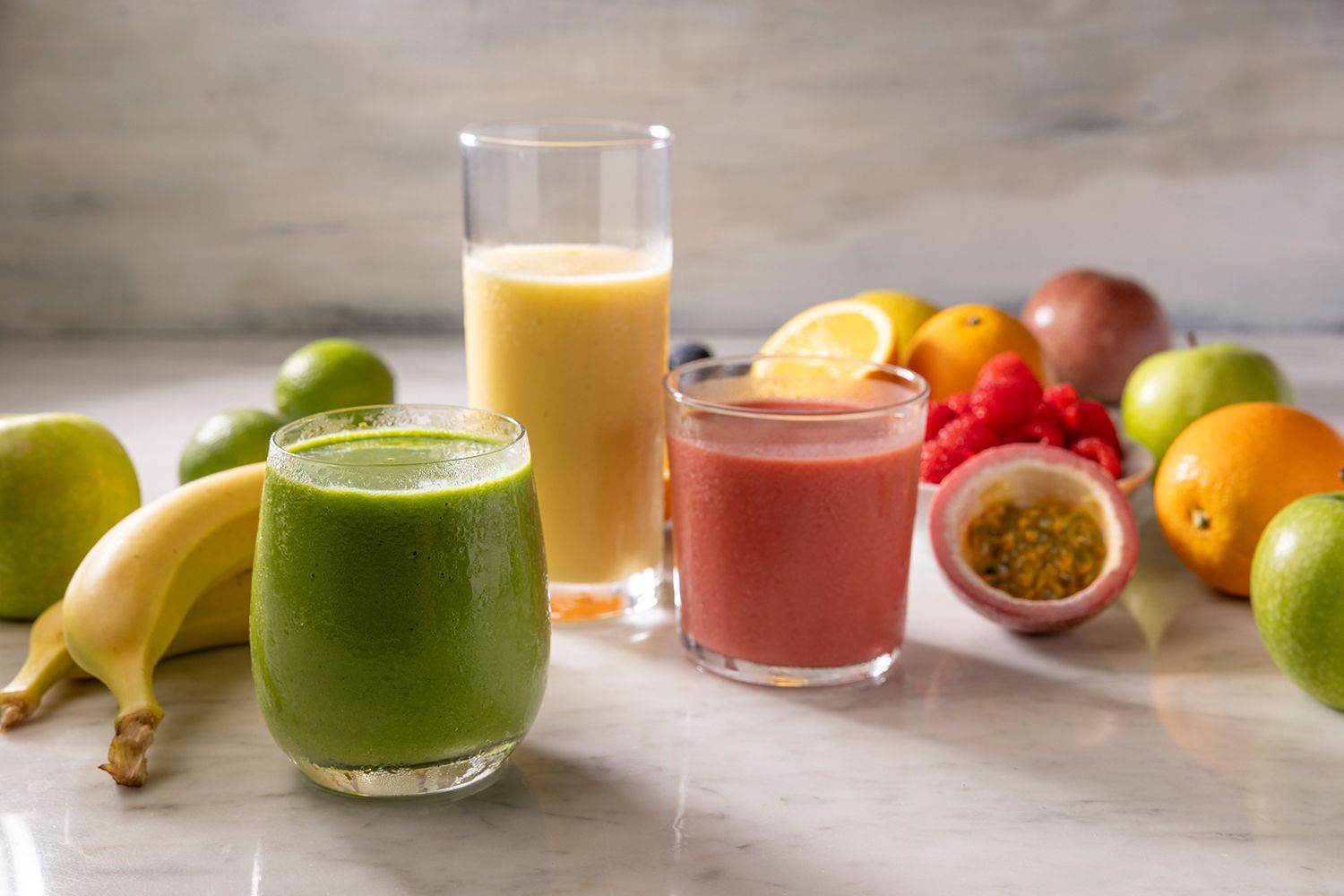 Three different smoothies are sitting on a table next to fruit.