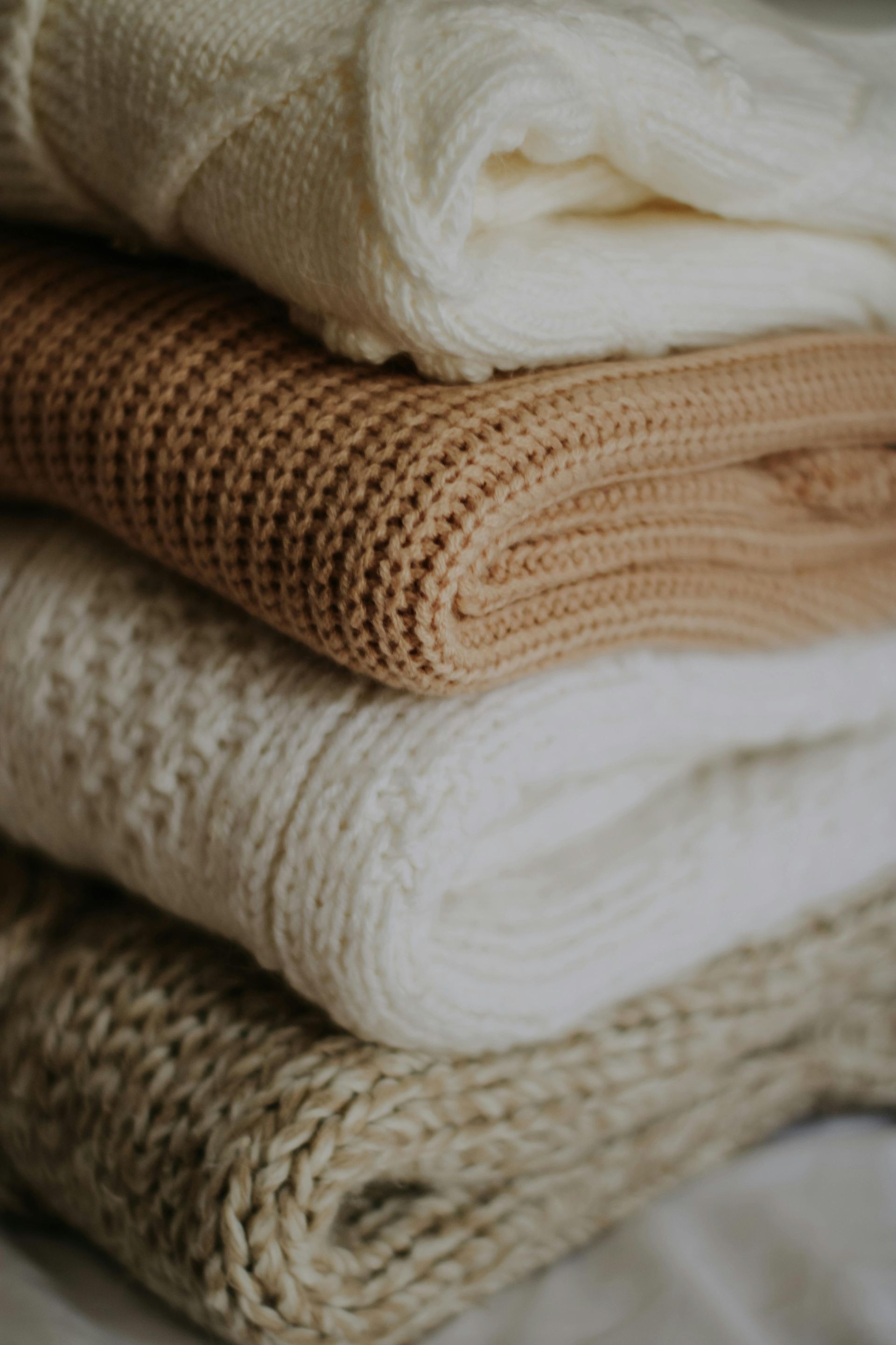 A stack of four folded knitted sweaters in various shades of cream, beige, and tan.