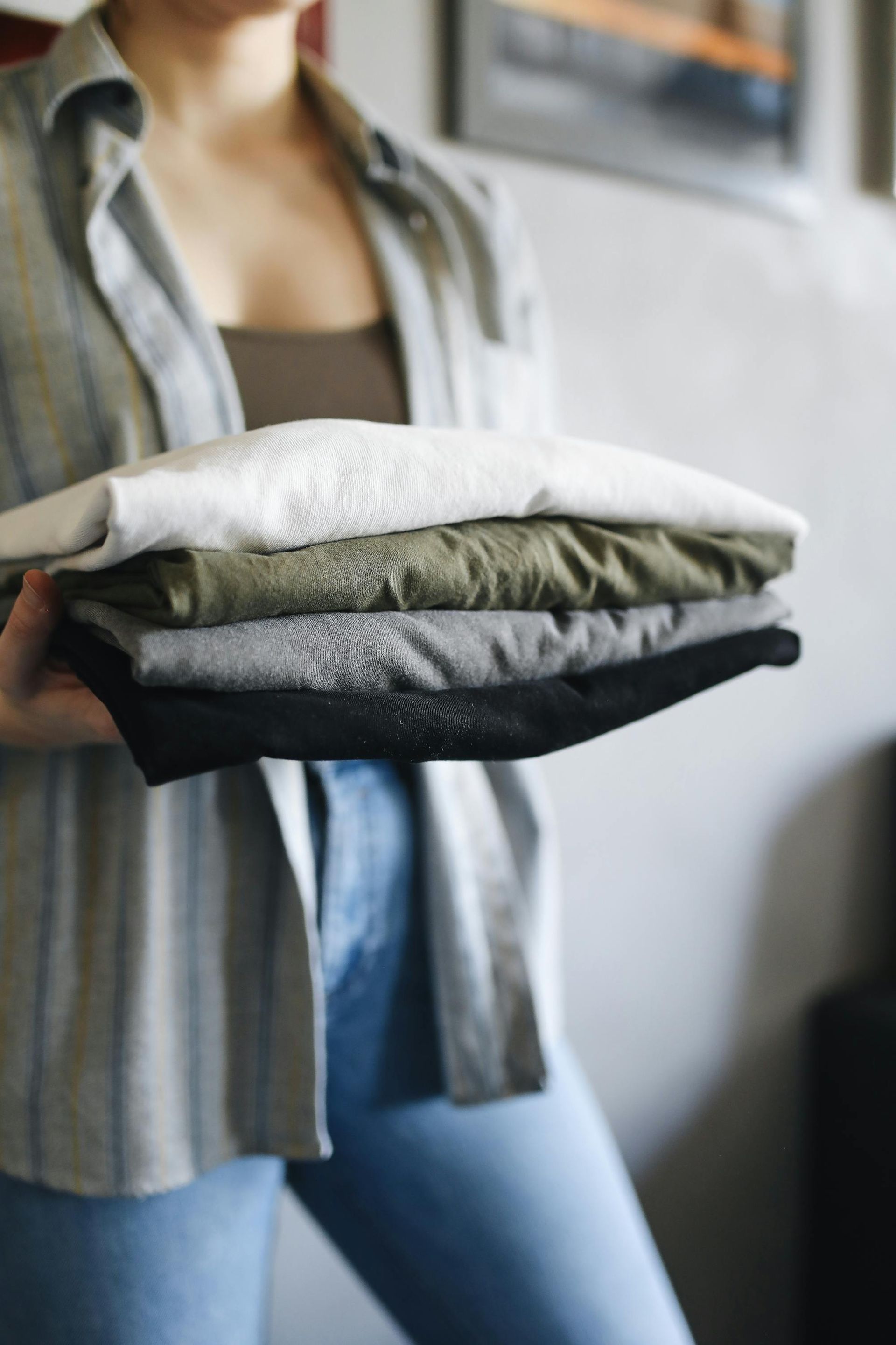 A person holds a neat stack of folded shirts in shades of white, olive, gray, and black.