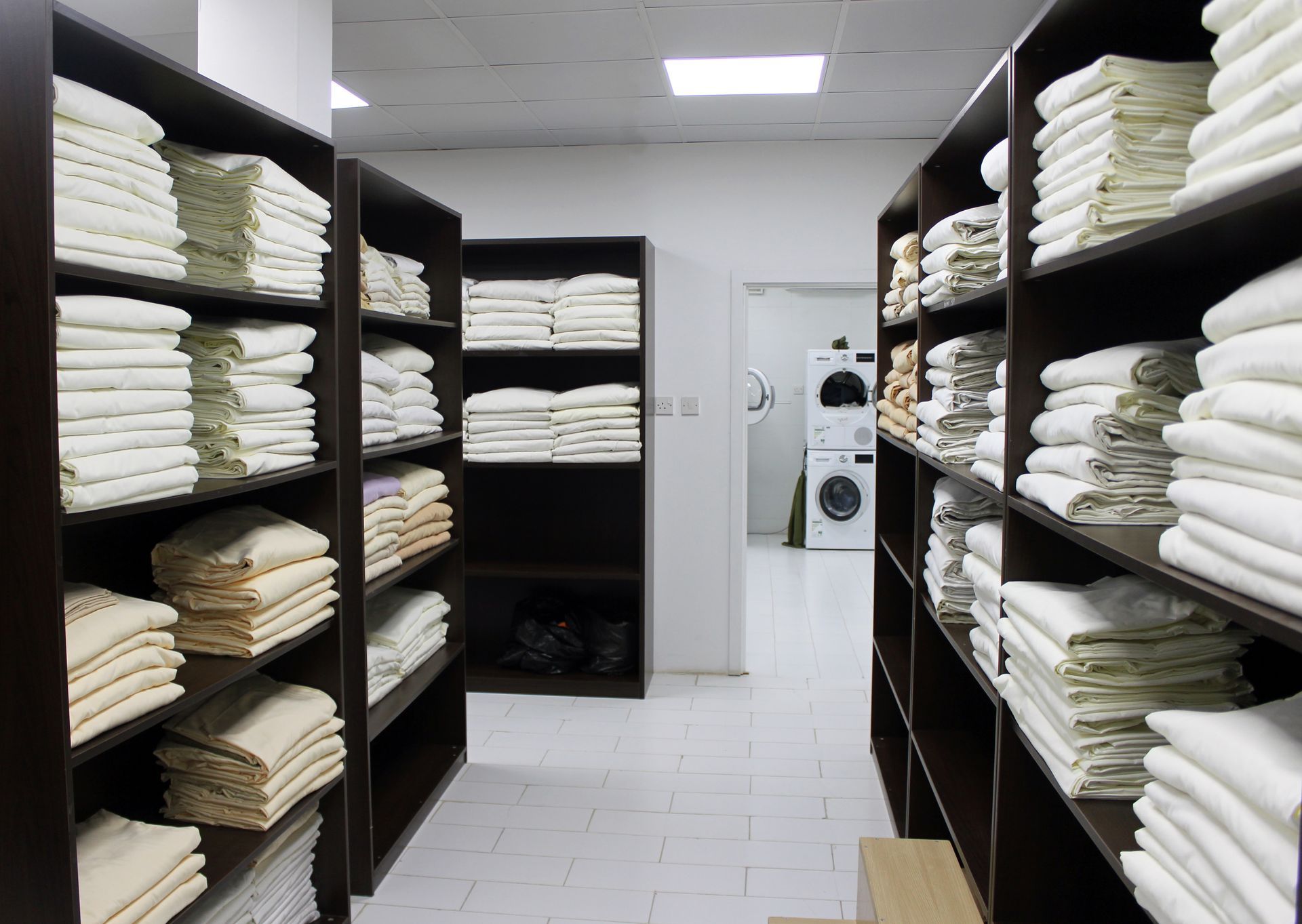 Rows of dark wooden shelves filled with neatly folded white linens in a brightly lit laundry storage room.