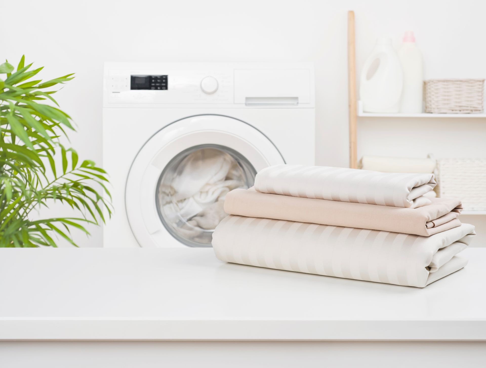 A white washing machine with a stack of folded laundry on a table in front of it, beside a potted plant and shelf.