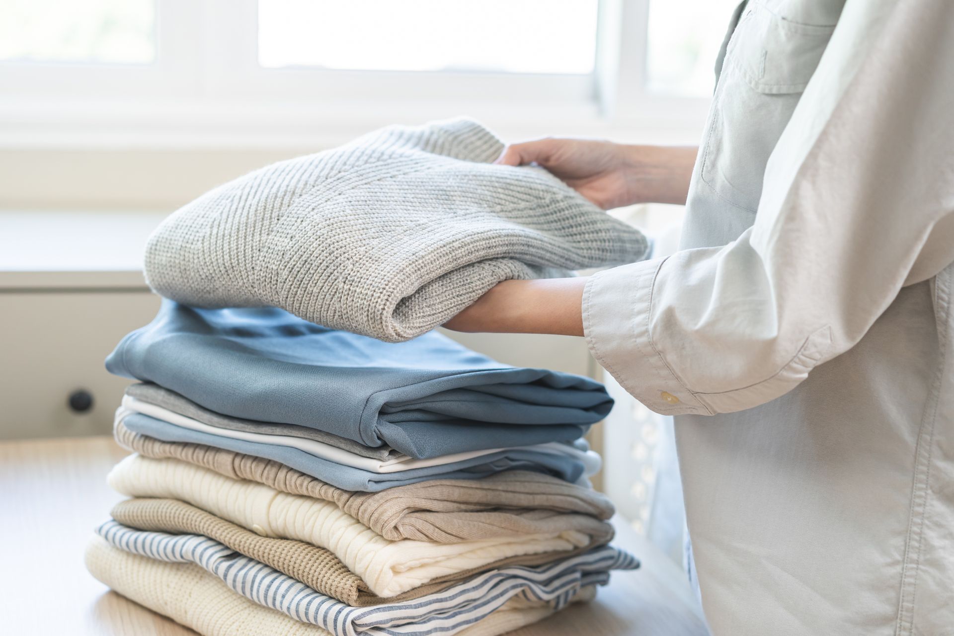 A person stacks folded, textured sweaters and garments in neutral colors on a table.