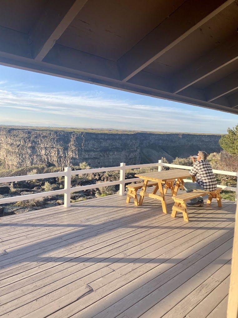 A man is sitting at a picnic table on a deck overlooking a canyon.