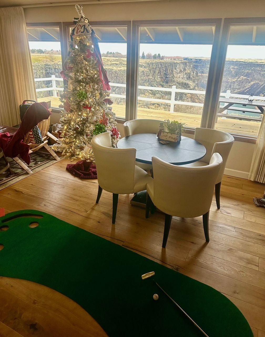 A Christmas tree stands near a dining table with chairs, overlooking a canyon. A golf putting green is in the foreground.