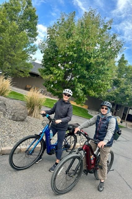 Two people on bikes posing outdoors. Woman in black, man in tan pants, green trees.