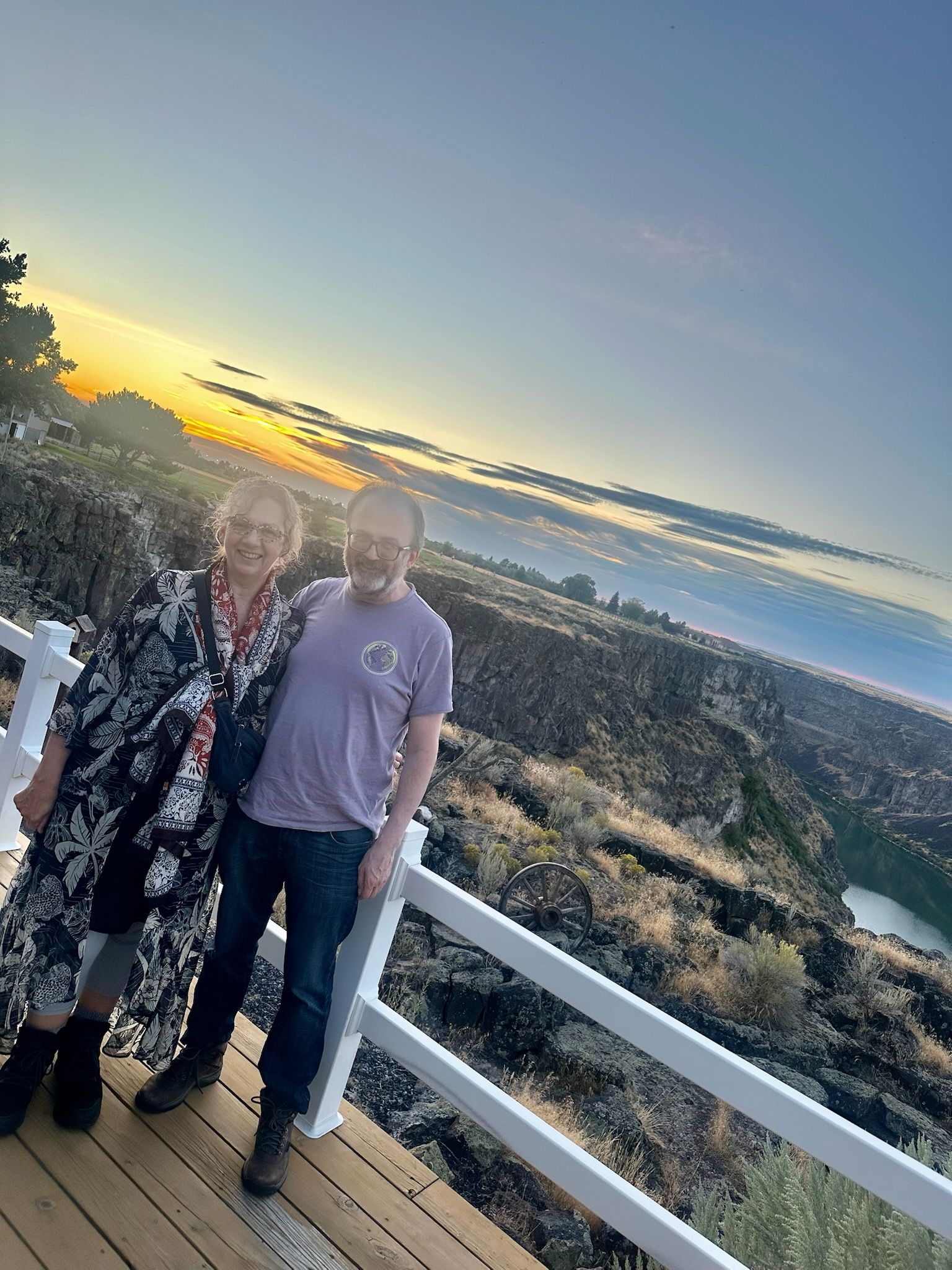 Two people smiling on a wooden deck, overlooking a canyon at sunset.