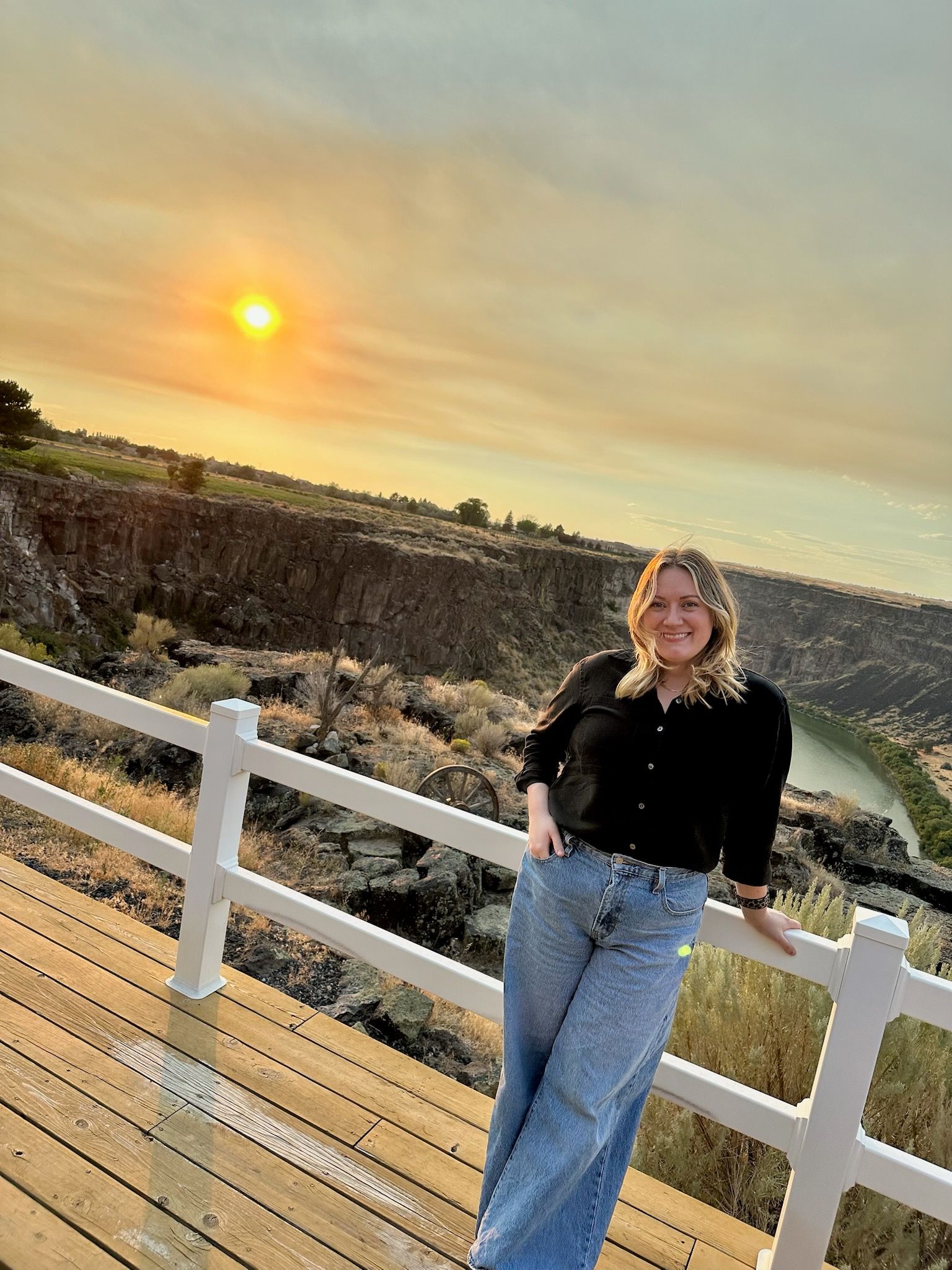 Woman on a deck overlooking a canyon at sunset. She wears a black shirt and jeans, smiles.