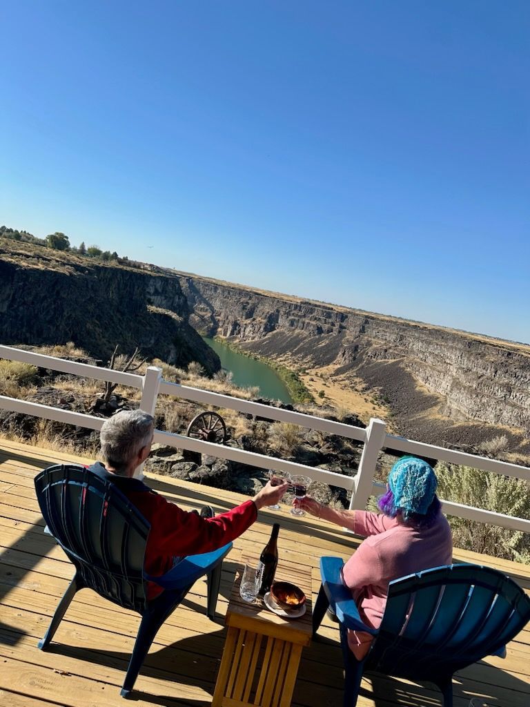 Two people toast drinks on a deck overlooking a canyon with a river.