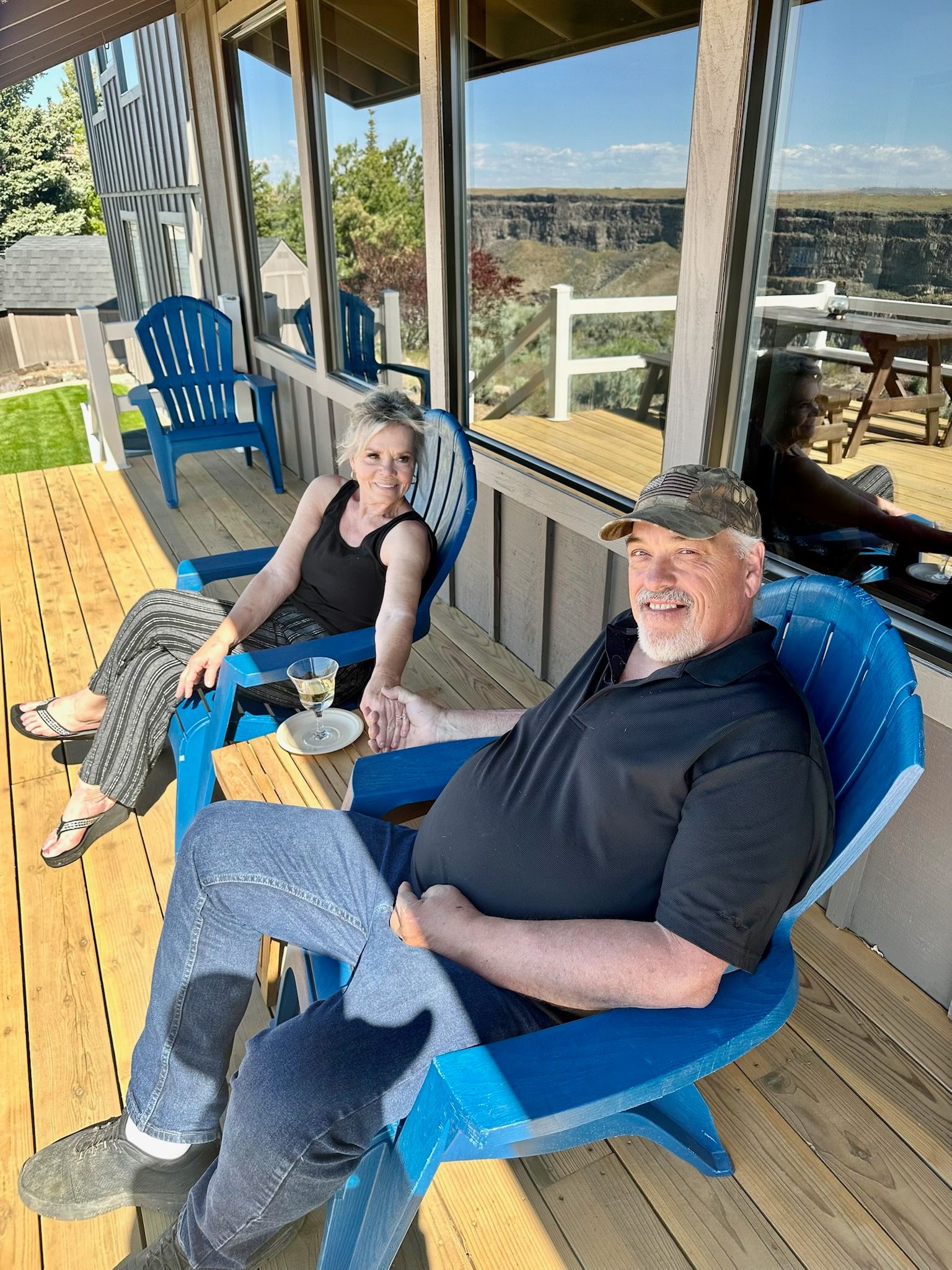 Couple holding hands, seated in blue Adirondack chairs on a porch, canyon view.