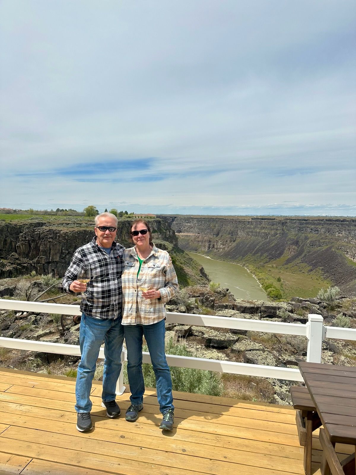 A couple poses on a wooden deck overlooking a canyon with a river; cloudy sky above.