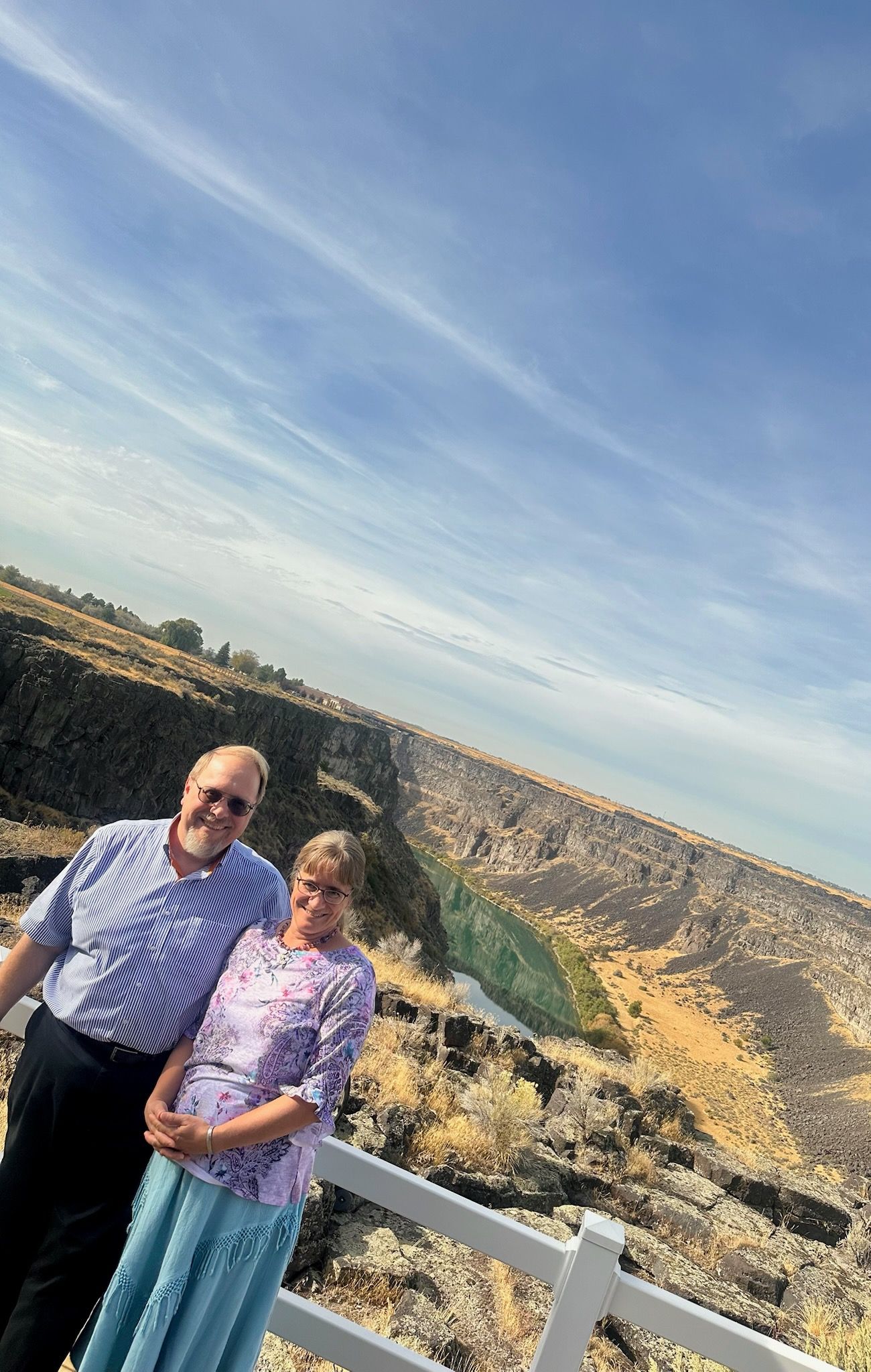 A couple smiles, posing on a cliff edge with blue sky above.