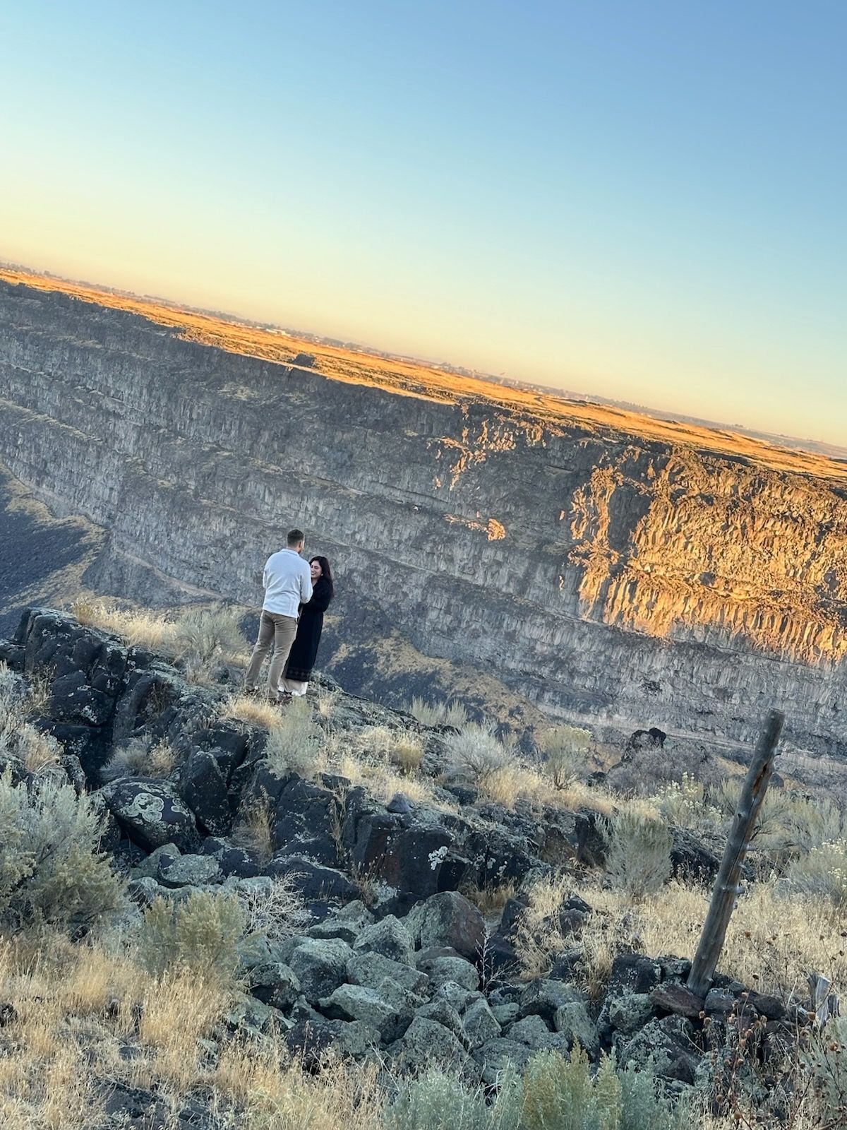 Couple standing on a rocky ridge, facing a canyon at sunset; golden light.