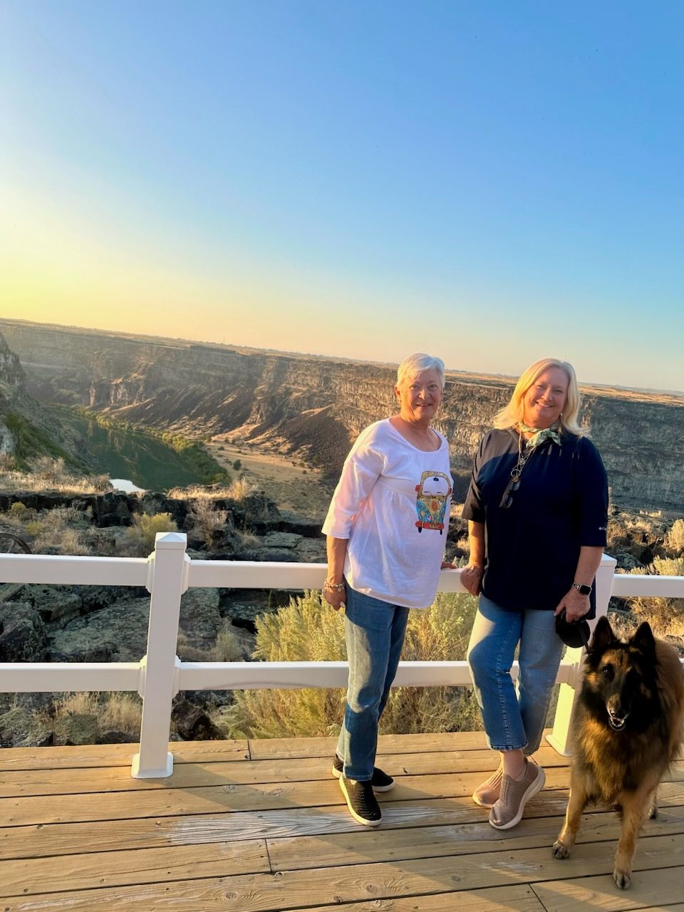 Two women and dog on a wooden deck overlooking a canyon at sunset.