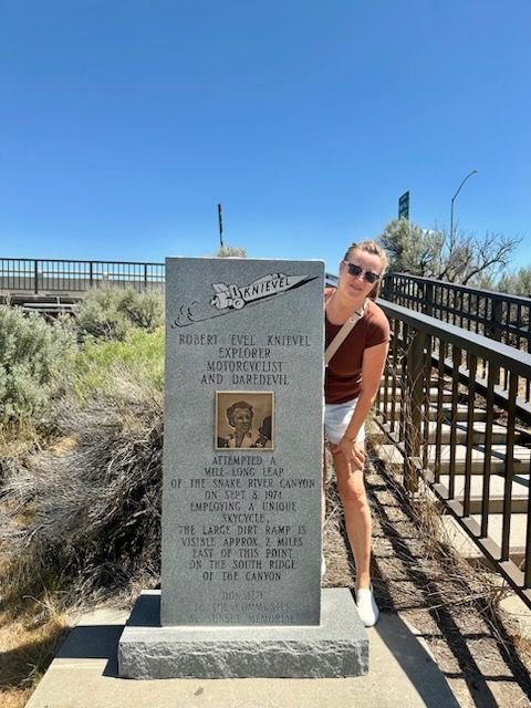 Woman beside a stone memorial with a portrait, brown top, white shorts, bridge and blue sky.