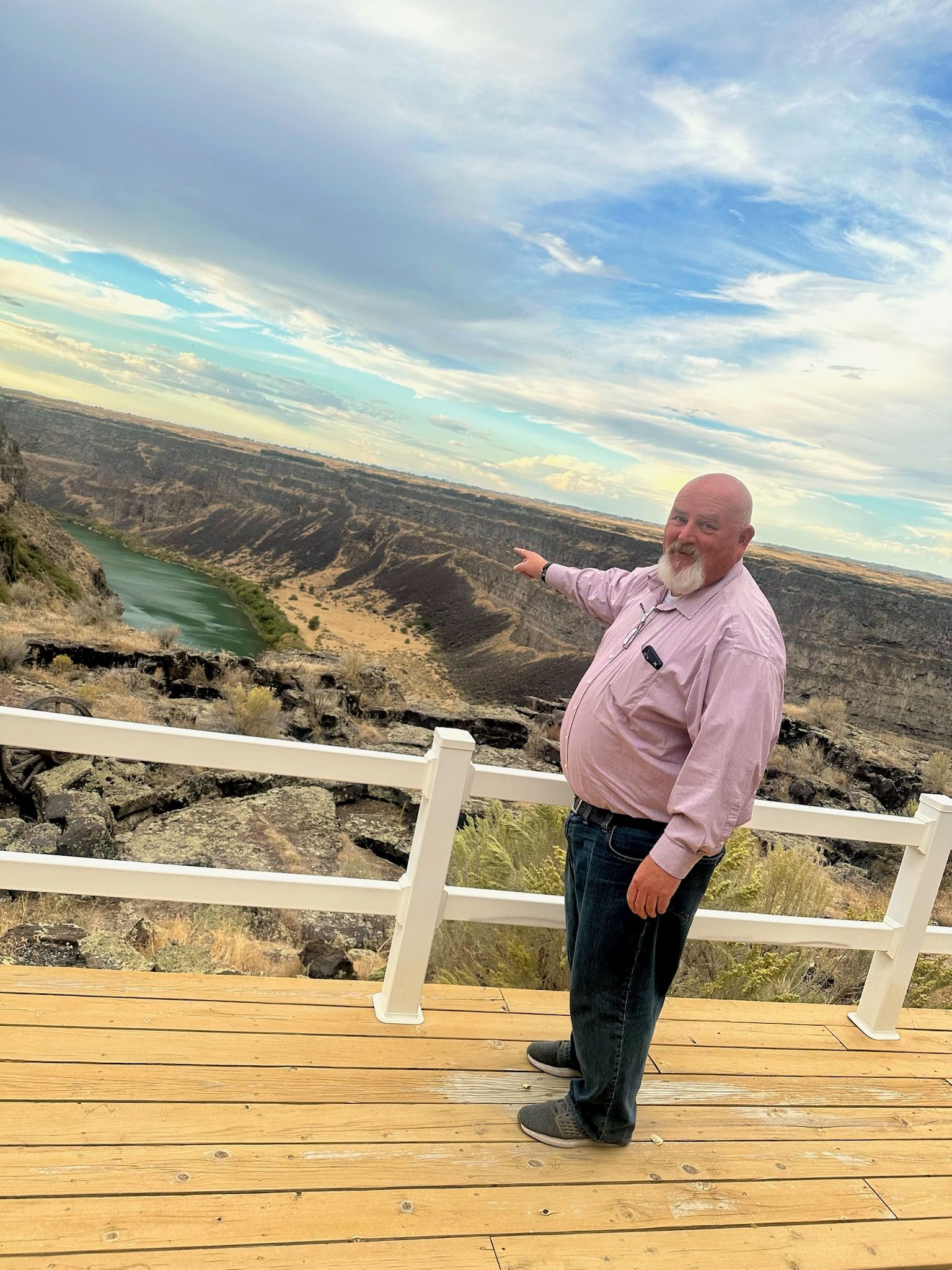 Man points at distant river flowing through a rocky landscape under a cloudy blue sky.