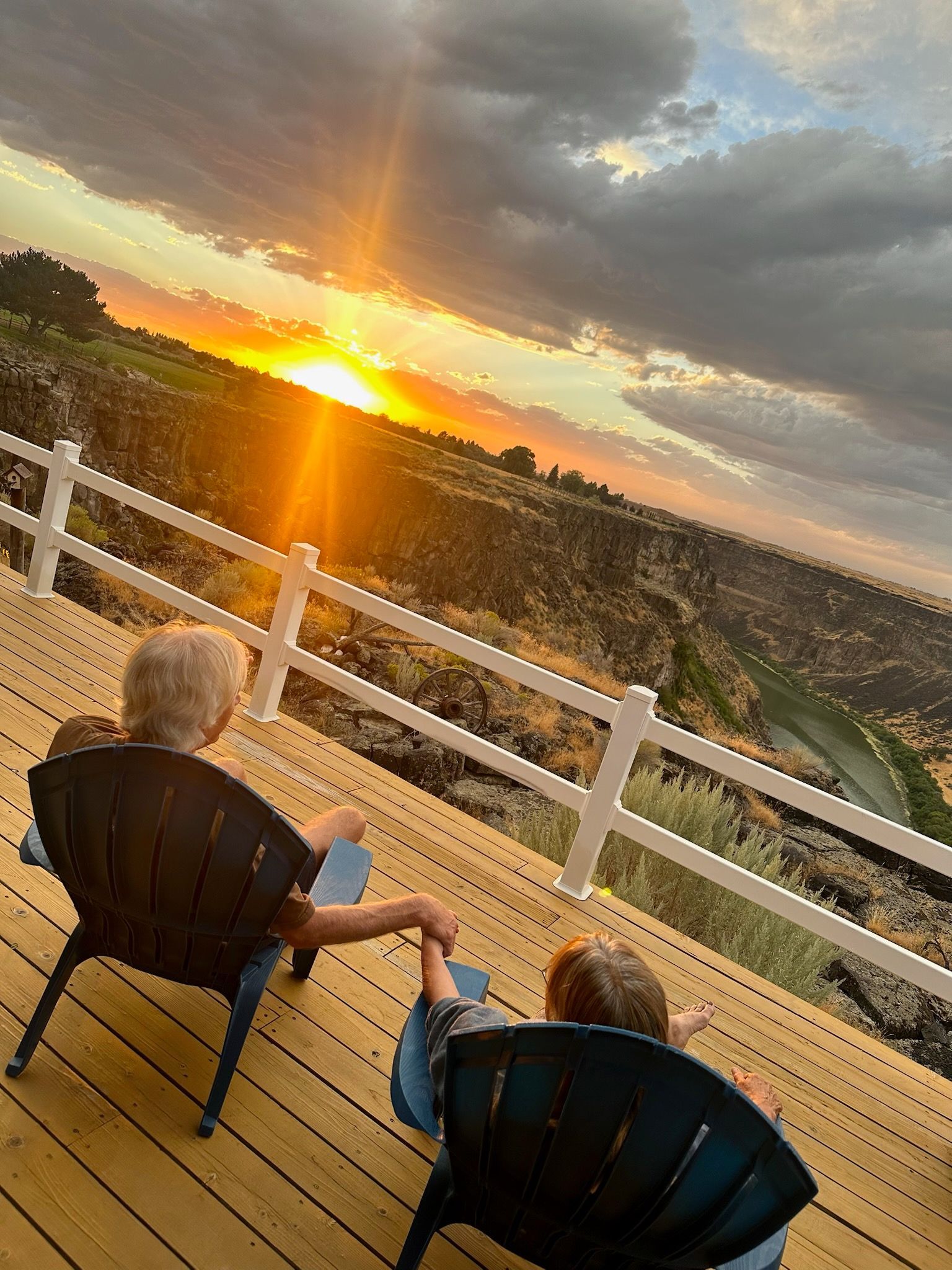 Two people holding hands on a deck watching a sunset over a scenic vista.