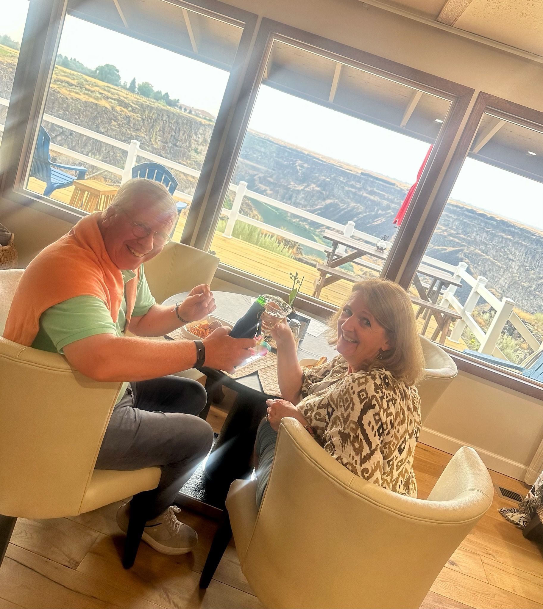 Couple sitting in chairs by a window, smiling and holding glasses. Overlooking a canyon.