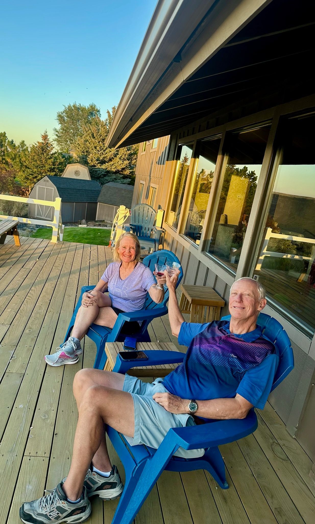 Two people sit in blue chairs on a wooden deck. Man holds a glass, smiling. Woman smiles too. Sunset in the background.