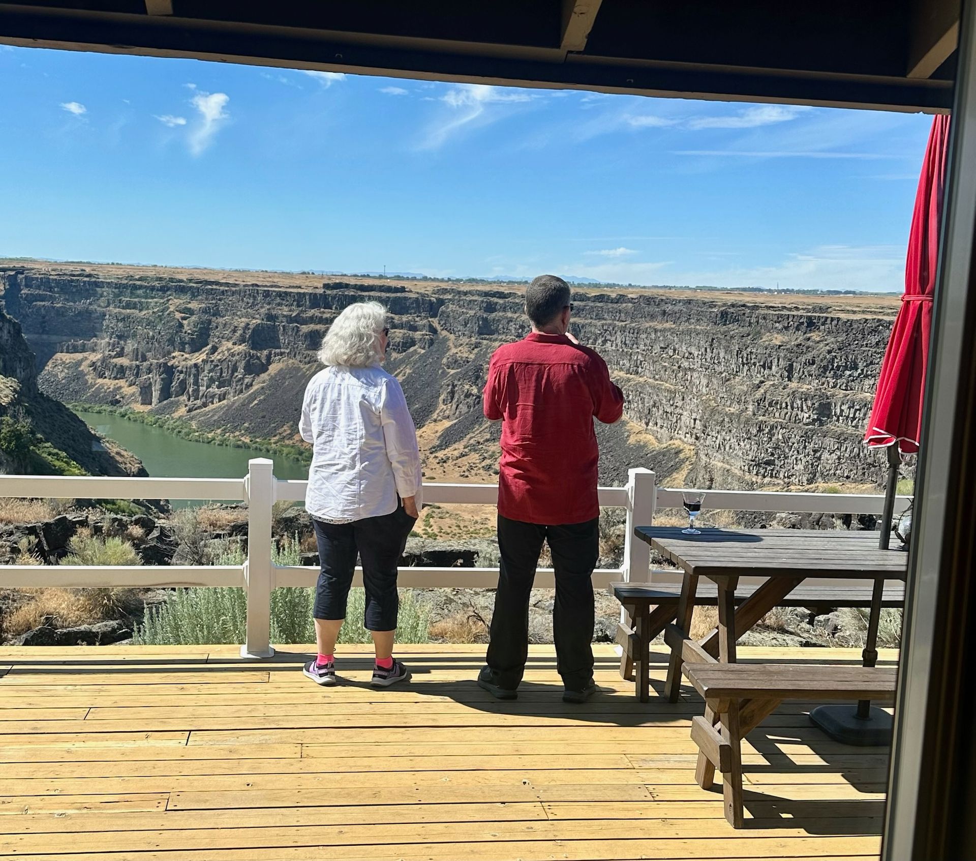 Two people on a deck overlooking a canyon, clear blue sky, a red umbrella, and a wood table.