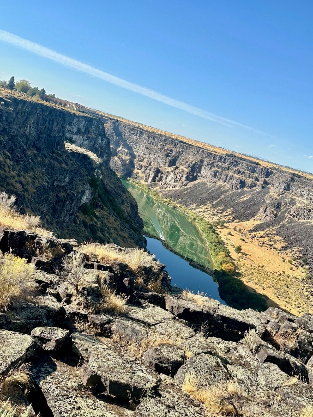 Canyon landscape with a blue river reflecting the sky, bordered by cliffs and dry vegetation under a clear sky.