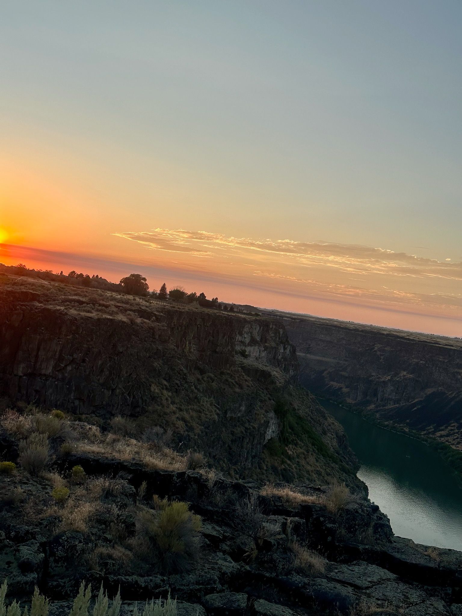 Sunset over a canyon with a dark, rocky cliff and water below. Orange sky.
