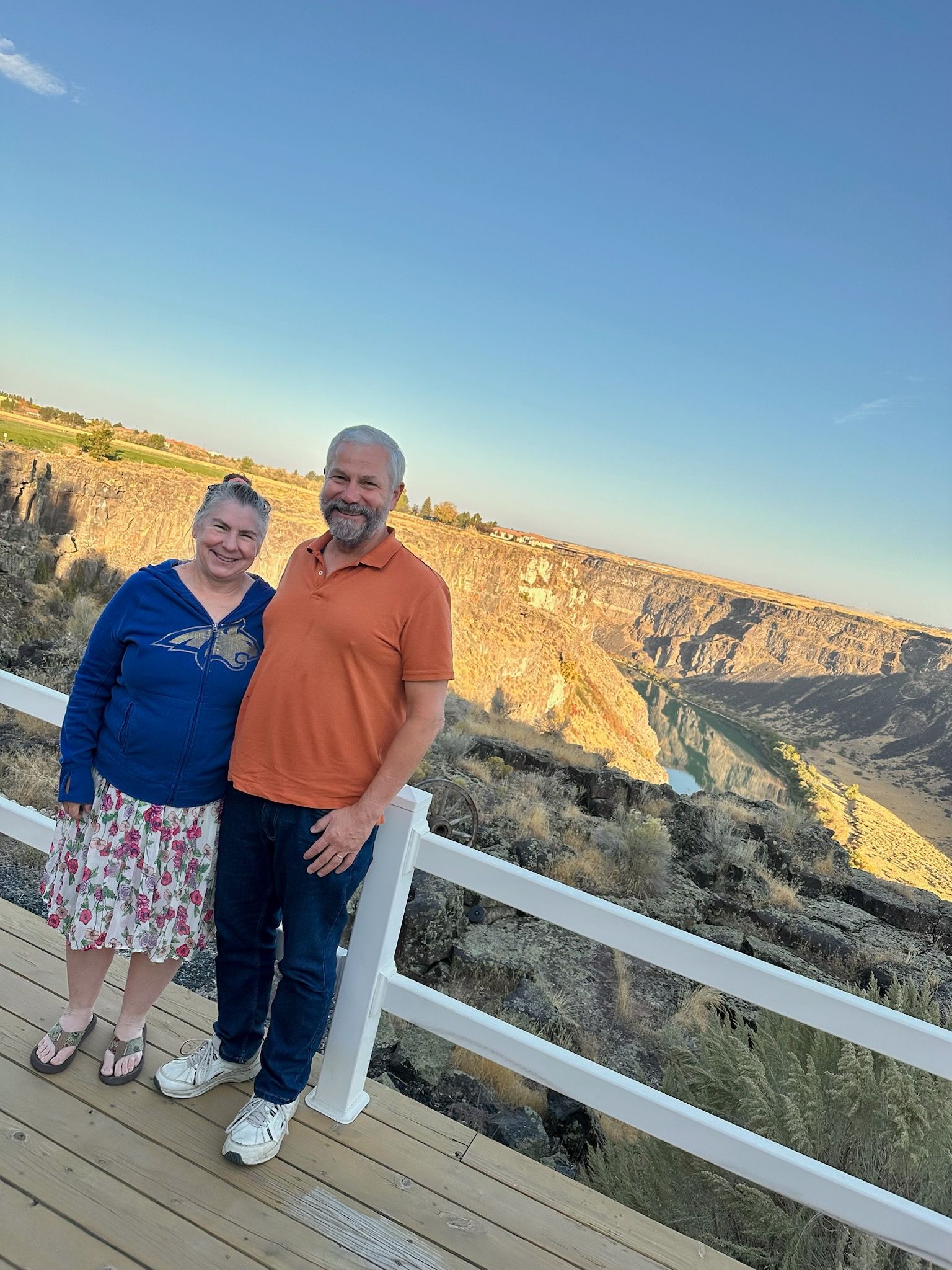 A couple poses for photo overlooking a canyon. The man wears orange, jeans, and the woman is in blue and floral skirt.