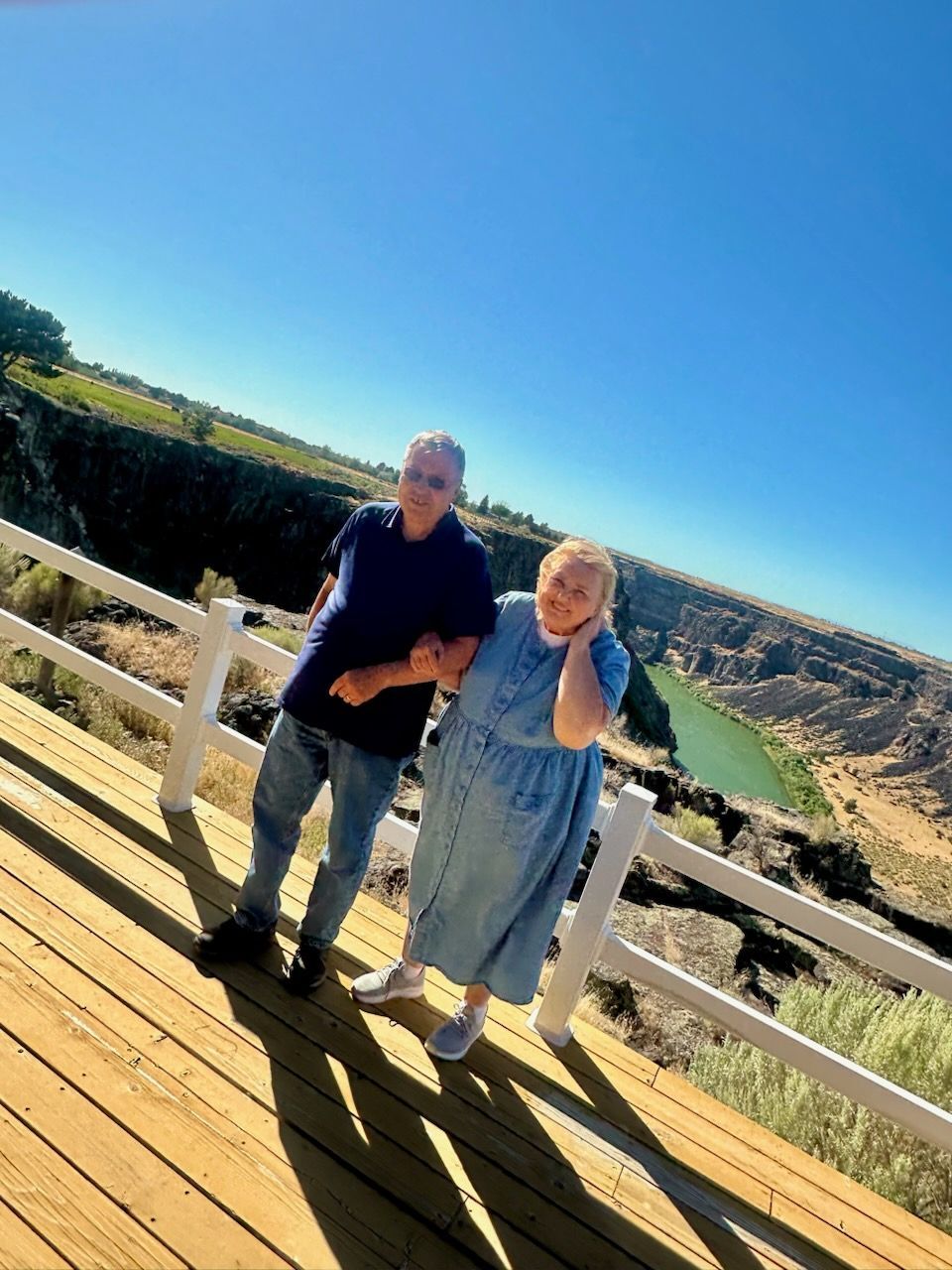 Two people, arm in arm, pose on a wooden deck overlooking a canyon with a river. Sunny day, blue sky.